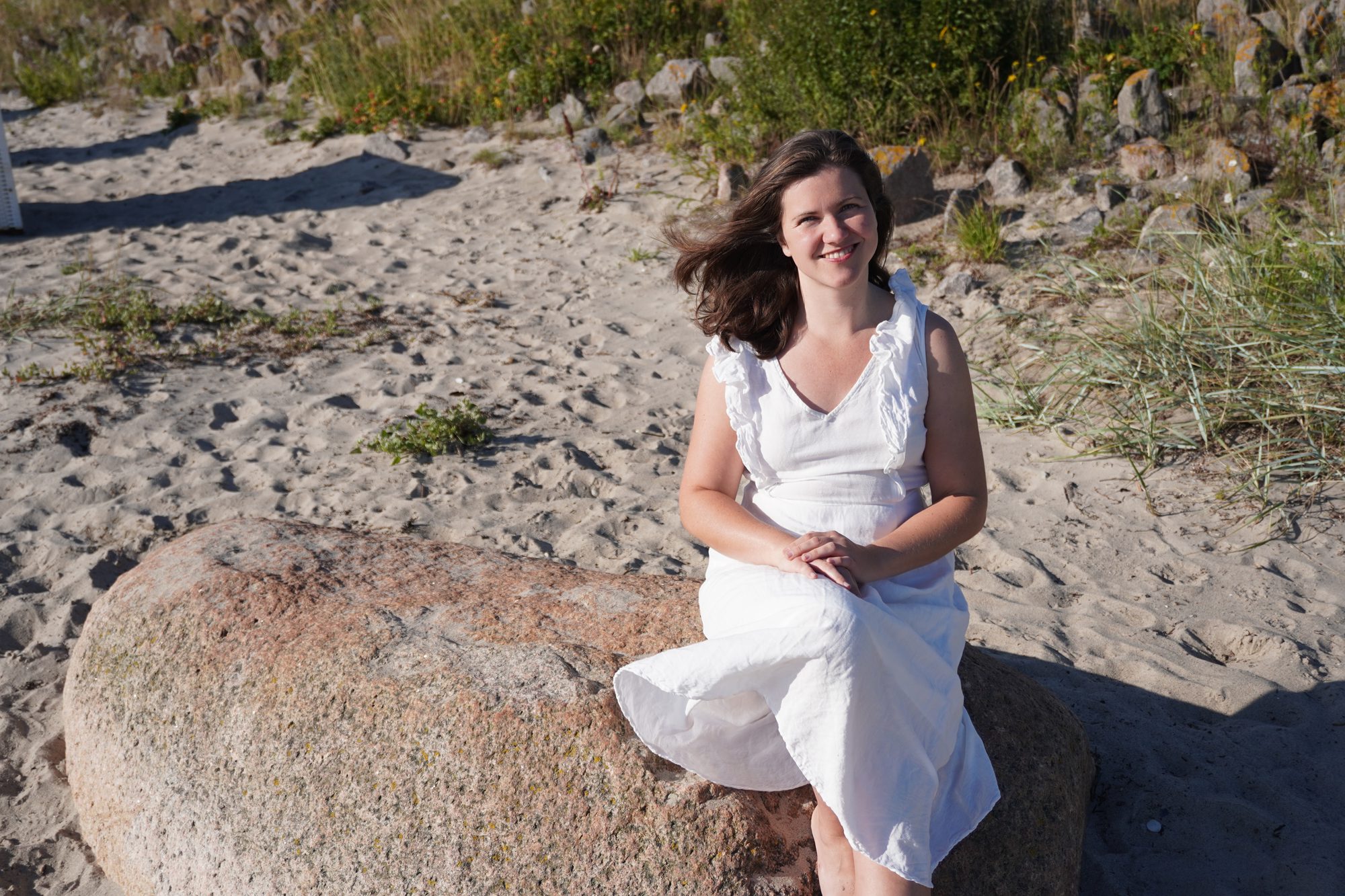 Rebecca Fackler sitzt im weißen Kleid auf einem Findling am Strand und schaut lächelnd in die Kamera