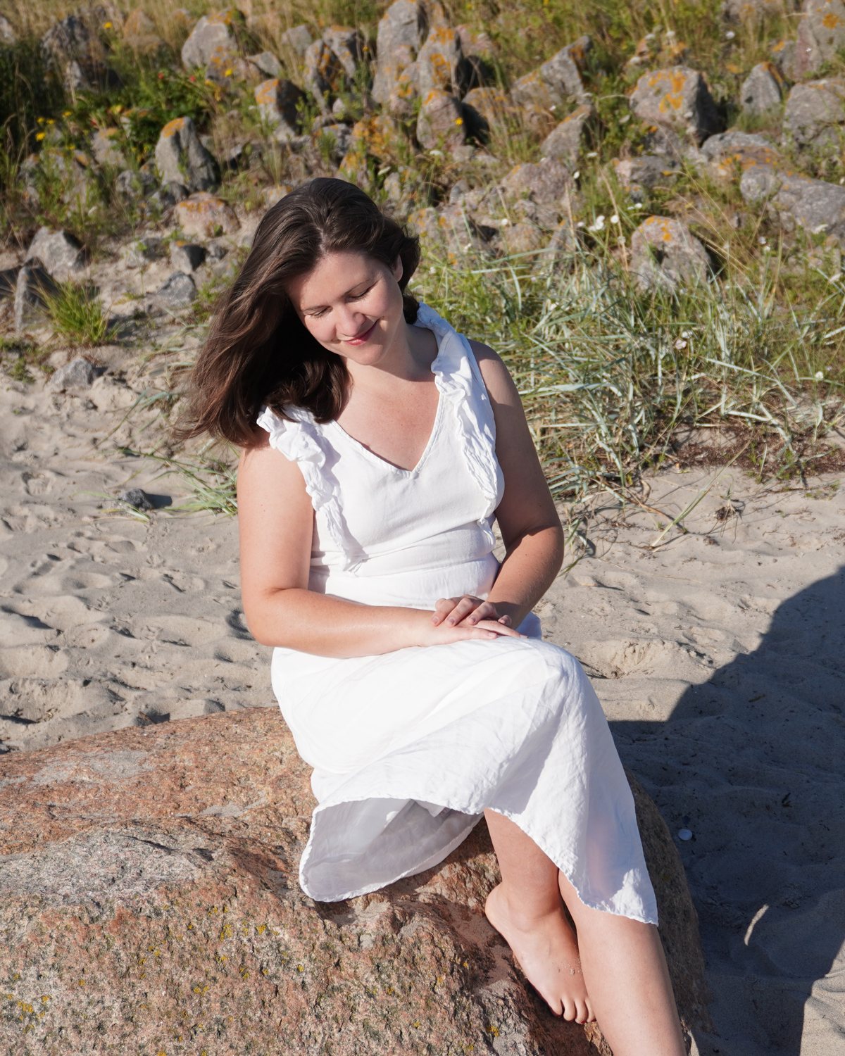 Rebecca Fackler sitzt im weißen Kleid auf einem Findling am Strand, den Blick nach unten gerichtet